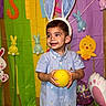 blue_outfit, bunny_ears, cartoon_chick, celebration, child, colorful_backdrop, cute, easter_theme, festive, happy, holiday, indoor, party_decorations, person, playful, plush_bunny, portrait, smiling, toddler, yellow_ball