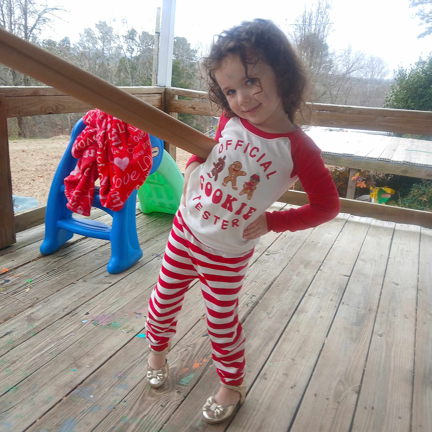 Willow is registered to the contest to win money with this photo: blanket, child, curly_hair, daylight, fence, footwear, gold_shoes, holiday_theme, outdoor, pajamas, person, play, playful, playset, posing, red_clothing, smiling, striped_clothing, trees, wooden_porch