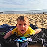 baby, child, beach, sand, towel, yellow_shirt, toy, ocean, sky, clouds, sunlight, happy, outdoor, infant, smiling, playful, water, summer, shore, vacation