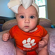 Violette is registered to the contest to win money with this photo: baby, infant, headband, bow, orange_onesie, cushion, purple, indoors, window, curious, face, blue_eyes, skin, hands, feet, soft_texture, person, child, portrait, cute