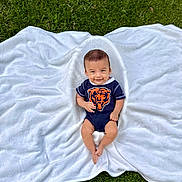 Mateo is registered to the contest to win money with this photo: baby, infant, smiling, onesie, bear_logo, blanket, grass, outdoor, cute, child, lying_down, greenery, happy, portrait, small, young, casual_clothing, nature, soft_texture, playful