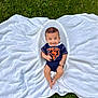 baby, infant, smiling, onesie, bear_logo, blanket, grass, outdoor, cute, child, lying_down, greenery, happy, portrait, small, young, casual_clothing, nature, soft_texture, playful