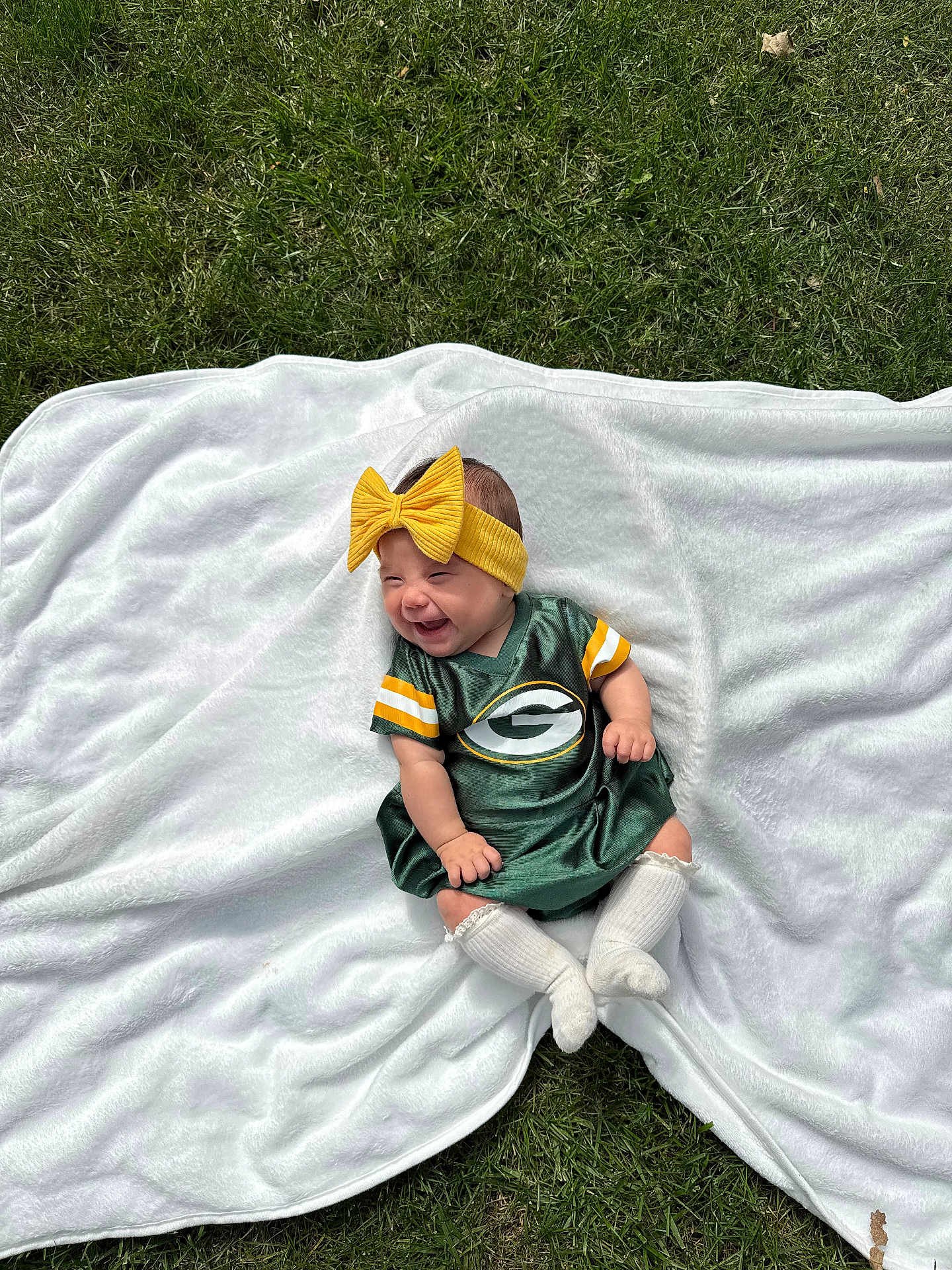 Luciella is registered to the contest to win money with this photo: baby, child, smiling, green_bay_packers, dress, yellow_bow, headband, white_blanket, grass, outdoor, happy, sitting, infant, cute, sock, white_sock, portrait, soft_texture, sportswear, baby_fashion