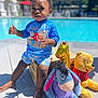 child, toddler, pool, outdoor, sunny, plush_toy, winnie_the_pooh, eeyore, balloon, blue_clothing, shorts, bracelet, barefoot, wooden_crate, concrete, summer, toy, person, expression, daylight
