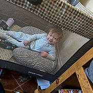 Matthias is registered to the contest to win money with this photo: baby, playpen, mesh, pillow, toy, floor, tile, child, smile, indoor, wooden_furniture, socks, white_clothing, happy, person, home, cozy, relaxed, infant, cushion
