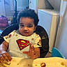 Noah joined the competition — help win amazing prizes! toddler, child, superman_logo, high_chair, food, crumbs, messy, curly_hair, kitchen, window, washing_machine, refrigerator, table, clothing, person, indoor, expression, face, hand, seat