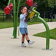 Aaliyah joined the competition — help win amazing prizes! child, playground, outdoor, greenery, flower, play_structure, concrete, grass, trees, fence, casual_clothing, sneakers, happy, curly_hair, summer, daytime, fun, kid, young, nature