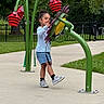 child, playground, outdoor, greenery, flower, play_structure, concrete, grass, trees, fence, casual_clothing, sneakers, happy, curly_hair, summer, daytime, fun, kid, young, nature