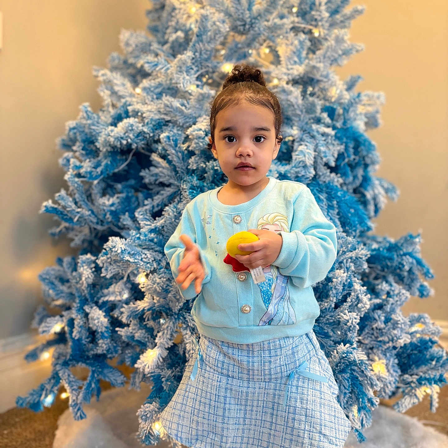 Aaliyah joined the competition — help win amazing prizes! toddler, child, christmas_tree, holiday, indoor, toy, blue, lights, frosted_tree, winter, cute, girl, clothing, skirt, sweater, white_tights, decorations, festive, portrait, home