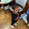 child, girl, lollipop, indoors, reading, card, black_clothing, pink_shoes, hair_bun, office, chair, table, papers, keyboard, floor, curious, standing, focused, toy, young