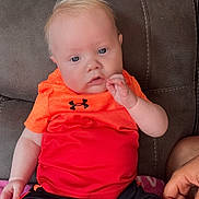 Tyde is registered to the contest to win money with this photo: baby, black_shorts, blonde_hair, casual_clothing, child, couch, curious_expression, cute, hand, indoor, infant, light_skin, orange_shirt, person, pink_blanket, portrait, seated, small_hand, support, under_armour