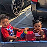 toddler, child, toy_wagon, parking_lot, car, sippy_cup, red_shirt, dress, sunlight, outdoor, curly_hair, playing, summer, cute, smiling, fun, two_children, casual, daytime, happy
