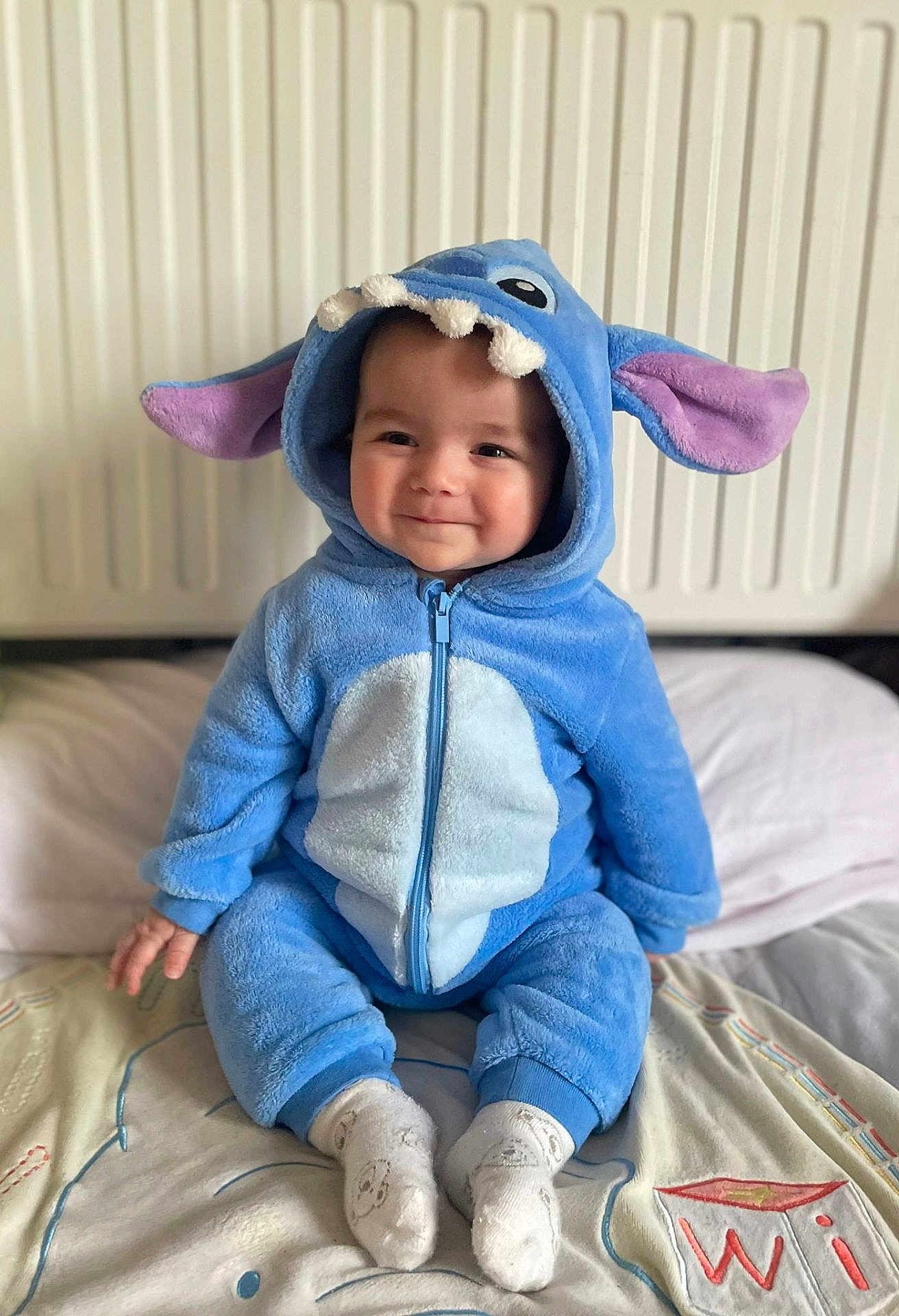Emilio a rejoint le concours — aidez-le/la à gagner de superbes lots ! baby, infant, child, blue_costume, onesie, hood, ears, smile, sitting, socks, bed, pillow, blanket, radiator, cozy, cute, portrait, face, zipper, soft_fabric