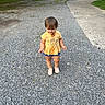 toddler, child, gravel, driveway, yellow_shirt, winnie_the_pooh, shorts, shoes, parked_cars, trees, house, outdoor, daylight, smiling, dirty_clothes, person, cute, young_child, standing, suburban