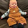 baby, child, tongue_out, overalls, brown, patterned_shirt, blue_socks, couch, leather, sitting, cute, playful, indoor, infant, smiling, person, small_hands, cozy, portrait, casual