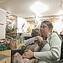 person, dog, indoor, christmas_tree, holiday_decorations, glasses, white_sweater, hugging, chair, gift_bags, living_room, warm_lighting, affection, pet, brown_dog, seated, furniture, cozy, home, casual