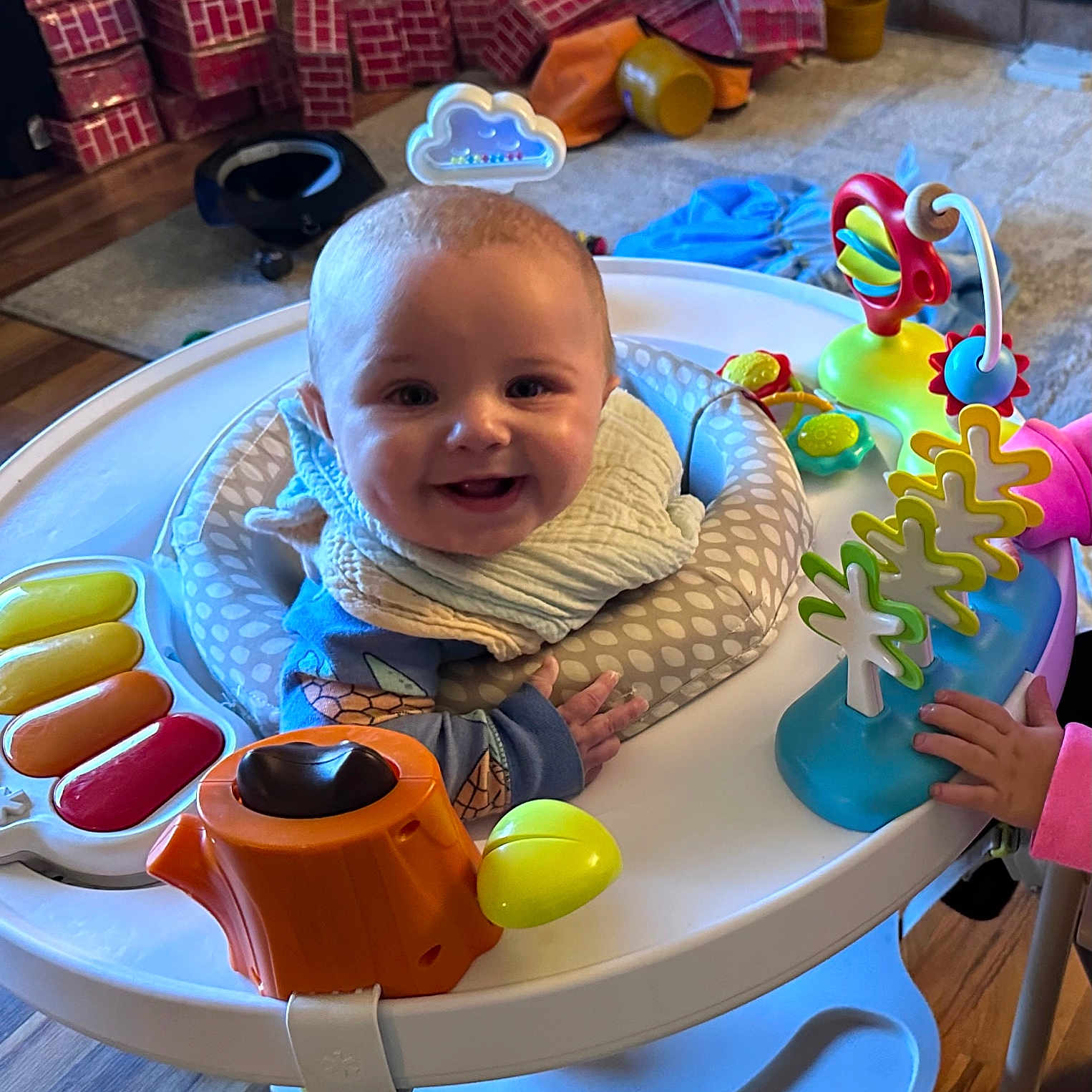 Maverick is registered to the contest to win money with this photo: activity_center, baby, bib, child, clothing, colorful, floor, furniture, hand, happy, indoor, infant, living_room, person, plastic, play, smiling, stacked_boxes, toy, wooden_floor