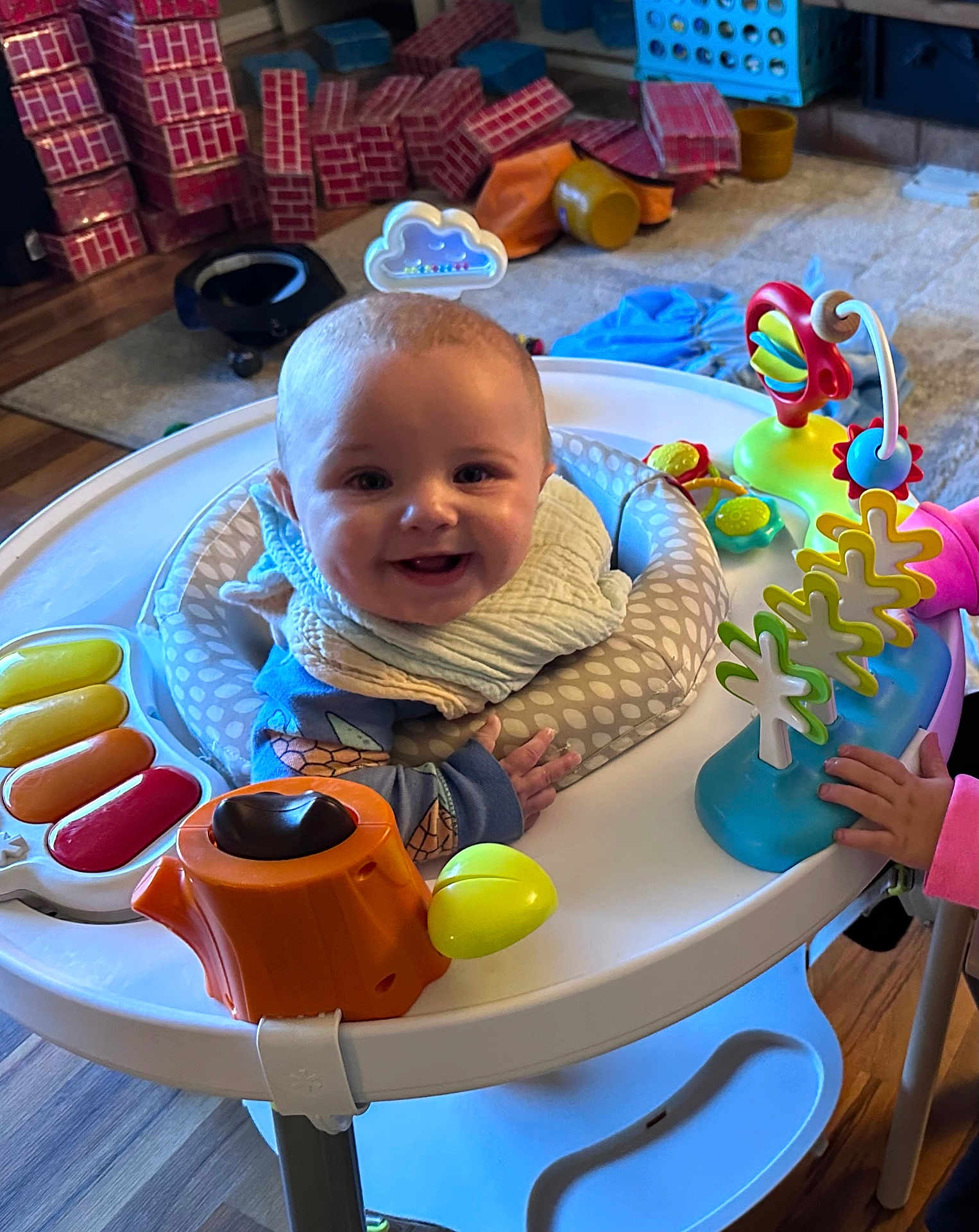 Maverick is registered to the contest to win money with this photo: baby, smiling, activity_center, bib, toy, hand, indoor, floor, wooden_floor, colorful, child, play, happy, person, clothing, furniture, plastic, infant, living_room, stacked_boxes