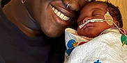 Riley is registered to the contest to win money with this photo: adult, baby, newborn, smiling, sleeping, blanket, headwrap, earring, nose_ring, hospital, medical_tube, close_up, face, skin, person, comfort, love, care, indoor, portrait