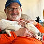 man, cat, indoor, smiling, beard, glasses, cap, orange_shirt, white_cat, gray_cat, sitting, couch, lamp, hand, relaxed, home, portrait, pet, comfort, happy