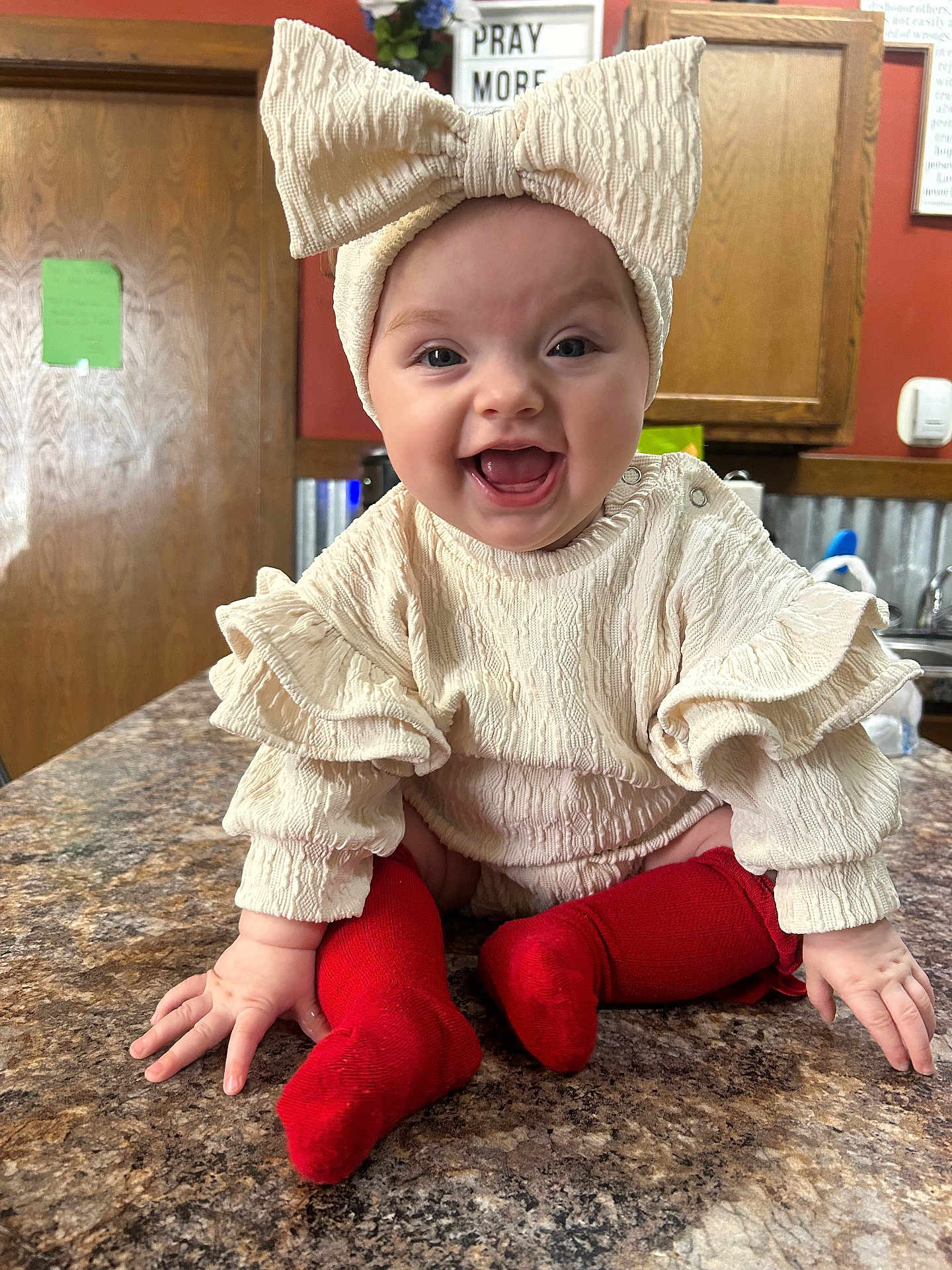 Emberlee joined the competition — help win amazing prizes! baby, infant, child, bow_headband, headband, smile, laughing, red_socks, ruffled_sleeves, countertop, kitchen, wooden_cabinet, door, portrait, indoor, sitting, happy, mouth_open, eyes, playful