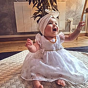 Lya participe au concours pour gagner de l'argent avec cette photo : baby, child, dress, white_dress, headwrap, smiling, happy, sitting, blanket, indoor, plant, wooden_floor, floor, arms_raised, cute, infant, portrait, cozy, decor, expression