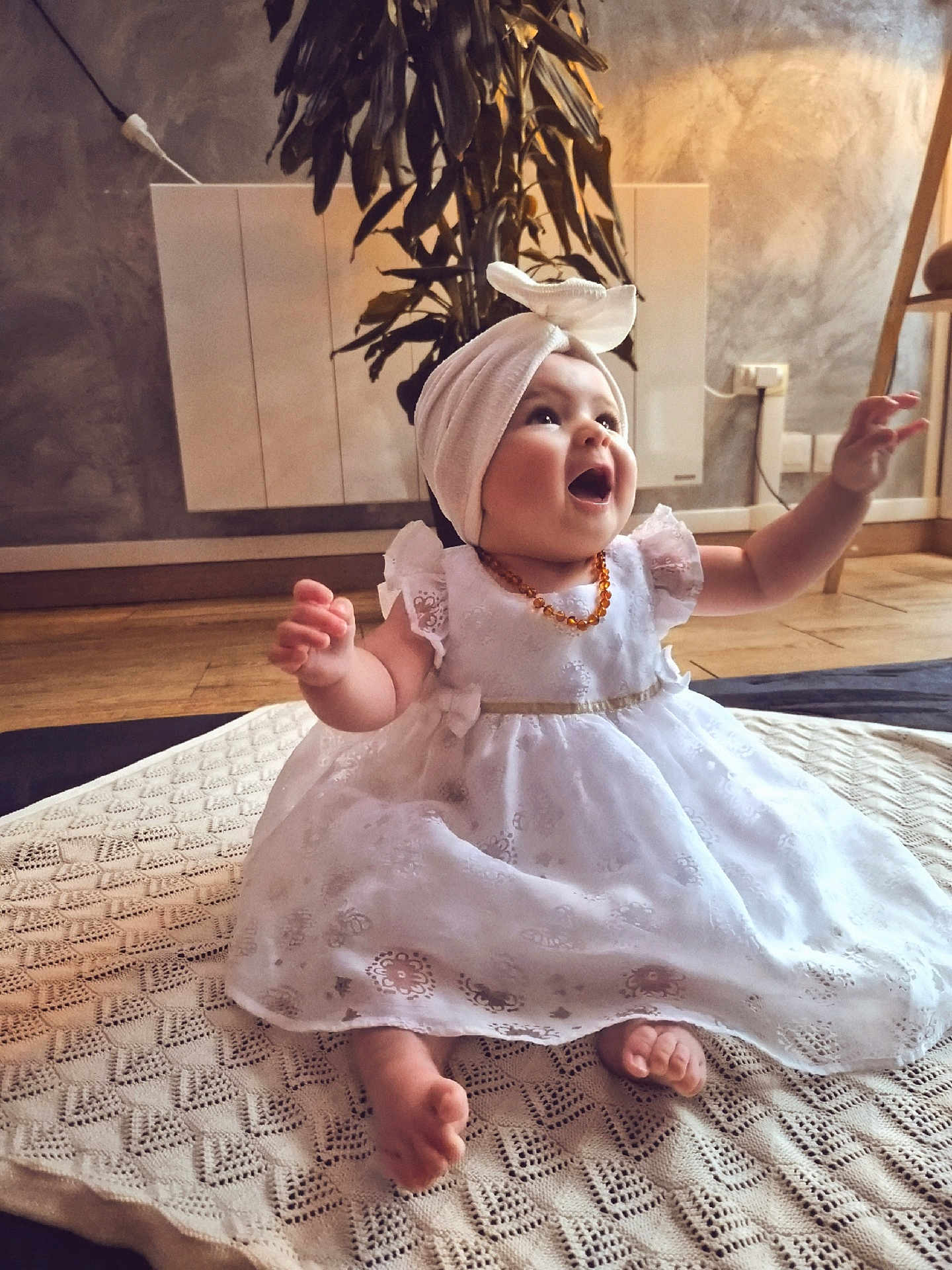 Lya participe au concours pour gagner de l'argent avec cette photo : baby, child, dress, white_dress, headwrap, smiling, happy, sitting, blanket, indoor, plant, wooden_floor, floor, arms_raised, cute, infant, portrait, cozy, decor, expression