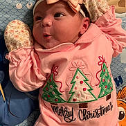 Rosalina is registered to the contest to win money with this photo: baby, infant, pink_clothing, headband, bow, christmas_theme, christmas_trees, patterned_blanket, cute, portrait, face, expression, lying_down, mittens, holiday, festive, child, soft_lighting, indoors, closeup