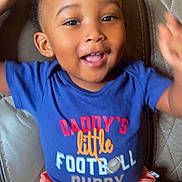 Messiah joined the competition — help win amazing prizes! child, toddler, smiling, happy, portrait, indoors, seated, leather_chair, blue_shirt, coral_shorts, football_shirt, casual_clothing, raised_arms, curly_hair, open_mouth, brown_eyes, living_room, close_up, happy_expression, photograph