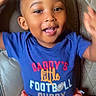 child, toddler, smiling, happy, portrait, indoors, seated, leather_chair, blue_shirt, coral_shorts, football_shirt, casual_clothing, raised_arms, curly_hair, open_mouth, brown_eyes, living_room, close_up, happy_expression, photograph