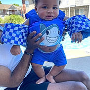 Kylen joined the competition — help win amazing prizes! baby, child, swimming_pool, floatation_device, blue, shark_design, adult_hand, outdoor, sunny, curly_hair, shorts, water_safety, summer, residential_area, person, skin, playing, happy, daytime, holding