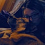 dog, person, glasses, indoor, dim_lighting, snuggle, pet, cozy, wood_paneling, window_blinds, couch, brown_dog, portrait, closeup, companion, relaxation, warmth, friendship, comfort, casual