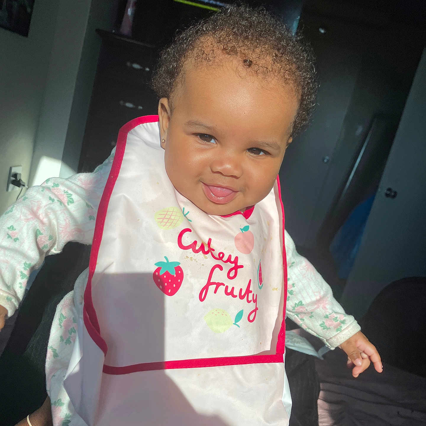 Kylani joined the competition — help win amazing prizes! baby, child, smiling, curly_hair, bib, indoor, light, shadow, clothing, face, person, cute, happy, portrait, toddler, furniture, television, room, sleepsuit, skin