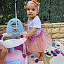 toddler, child, pink_tutu, headband, ride_on_toy, play_mat, toys, outdoor, greenery, stone_wall, barefoot, cute, playtime, colorful, happy, girl, summer, daylight, fashion, fun