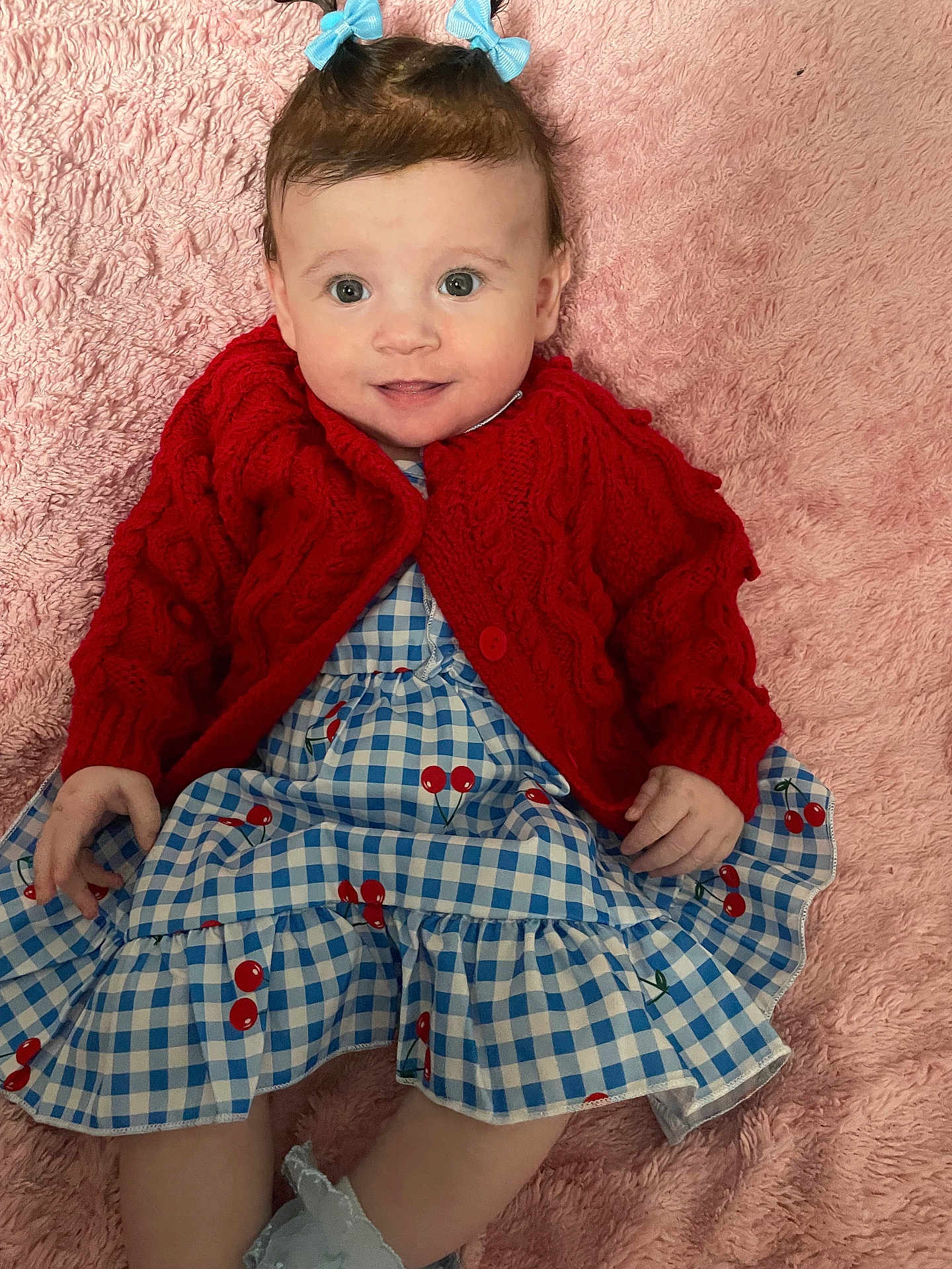 Payton is registered to the contest to win money with this photo: baby, child, smiling, blue_bows, red_cardigan, blue_gingham_dress, cherry_pattern, pink_blanket, soft_texture, cute, happy, portrait, infant, clothing, hands, legs, indoor, cozy, playful, innocence