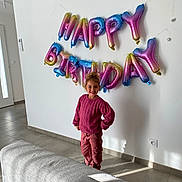 énora a rejoint le concours — aidez-le/la à gagner de superbes lots ! child, birthday_balloons, happy_birthday, pink_sweater, pink_pants, smiling, indoor, living_room, couch, tile_floor, air_conditioner, wall, door, window_light, curly_hair, standing, celebration, home, shadow, electric_socket