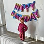 child, birthday_balloons, happy_birthday, pink_sweater, pink_pants, smiling, indoor, living_room, couch, tile_floor, air_conditioner, wall, door, window_light, curly_hair, standing, celebration, home, shadow, electric_socket