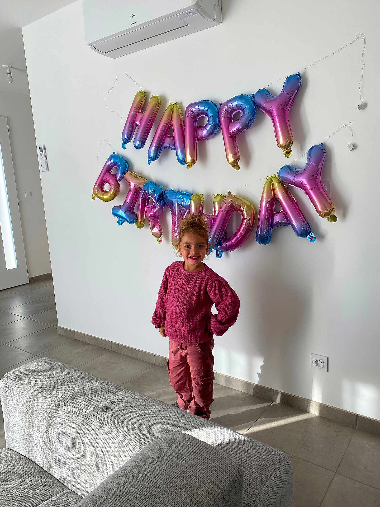 énora a rejoint le concours — aidez-le/la à gagner de superbes lots ! child, birthday_balloons, happy_birthday, pink_sweater, pink_pants, smiling, indoor, living_room, couch, tile_floor, air_conditioner, wall, door, window_light, curly_hair, standing, celebration, home, shadow, electric_socket