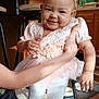 baby, smiling_baby, headband, bow, pink_dress, floral_applique, tutu, white_tights, ruffled_shoes, adult_hand, chair, stool, kitchen, cabinets, table_runner, carpet, floor, snack_bowl, hoodie, portrait