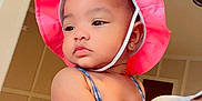 Malani is registered to the contest to win money with this photo: baby, child, pink_hat, blue_outfit, earring, indoor, person, adult_hand, holding, portrait, cute, chubby_cheeks, contemplative, clothing, skin, face, headwear, sleeveless, fashion, human