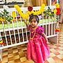 celebration, child, crib, curious, decor, dress, floor_tiles, flower_garland, furniture, gold_necklace, hair_accessory, indoor, jewelry, person, portrait, room, standing, toddler, traditional_clothing, young_girl