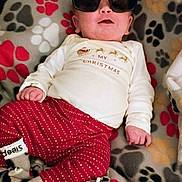 Jayce is registered to the contest to win money with this photo: baby, sunglasses, blanket, paw_prints, red_pants, christmas_shirt, infant, cozy, cute, sleep_socks, lying_down, indoors, festive, clothing, child, portrait, relaxed, home, fashion, funny