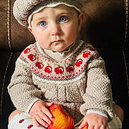 Aurora is registered to the contest to win money with this photo: toddler, child, knitted_hat, pom_pom, blue_eyes, red_apples, knitted_sweater, patterned_socks, sitting, brown_chair, indoor, cute, portrait, baby, cozy_clothing, curious_expression, hands, face, soft_lighting, warm_colors