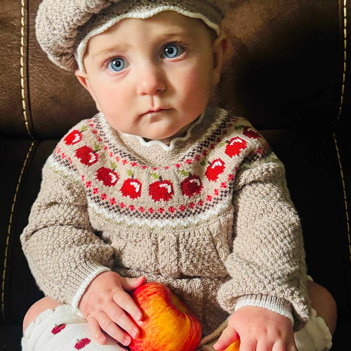 Aurora is registered to the contest to win money with this photo: toddler, child, knitted_hat, pom_pom, blue_eyes, red_apples, knitted_sweater, patterned_socks, sitting, brown_chair, indoor, cute, portrait, baby, cozy_clothing, curious_expression, hands, face, soft_lighting, warm_colors
