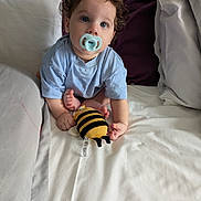 Leo joined the competition — help win amazing prizes! baby, child, pacifier, curly_hair, blue_eyes, plush_toy, bee_toy, bed, pillow, blanket, clothing, infant, cute, sitting, indoors, soft_light, person, young_child, cozy, comfort