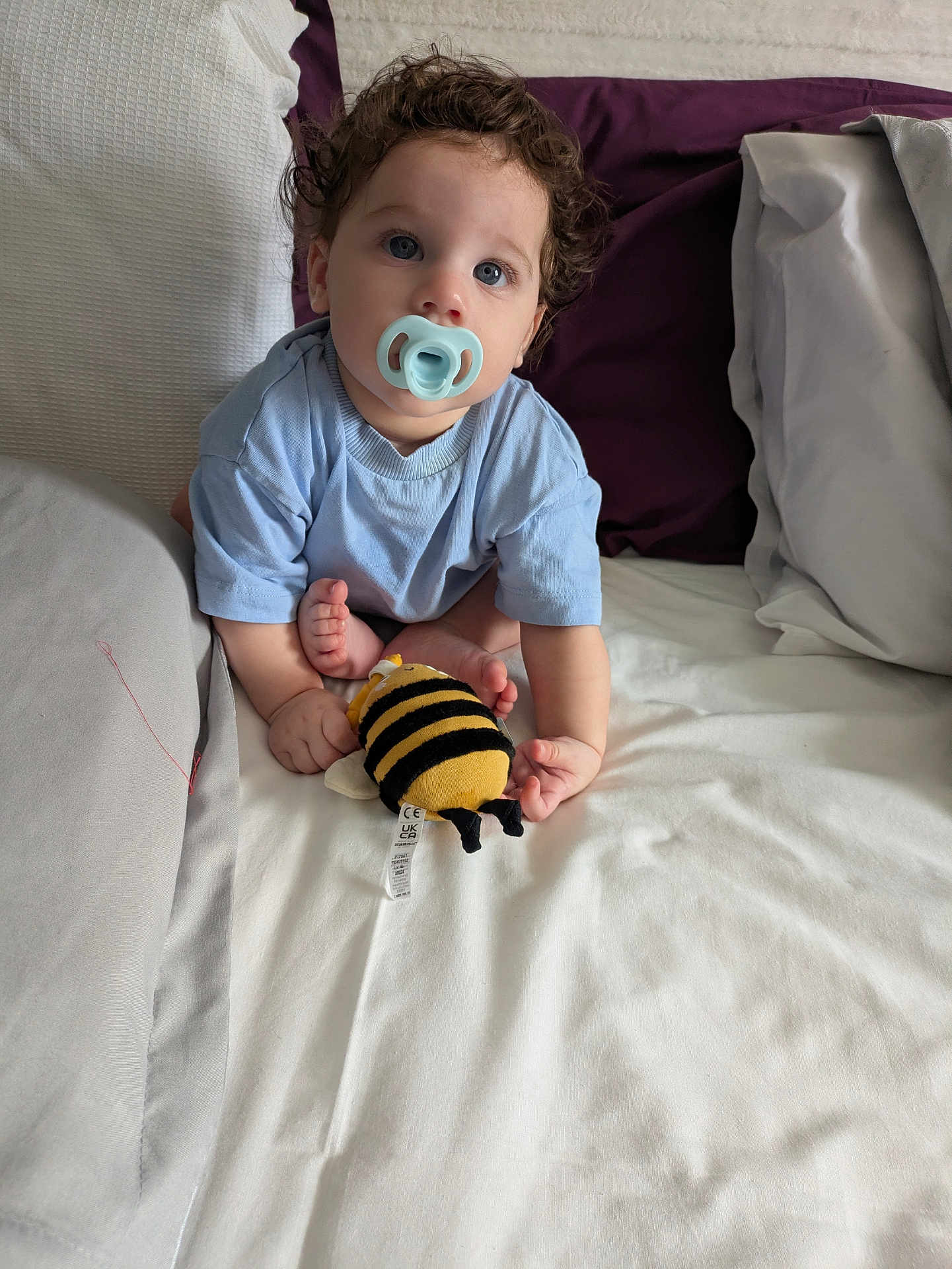 Leo joined the competition — help win amazing prizes! baby, child, pacifier, curly_hair, blue_eyes, plush_toy, bee_toy, bed, pillow, blanket, clothing, infant, cute, sitting, indoors, soft_light, person, young_child, cozy, comfort