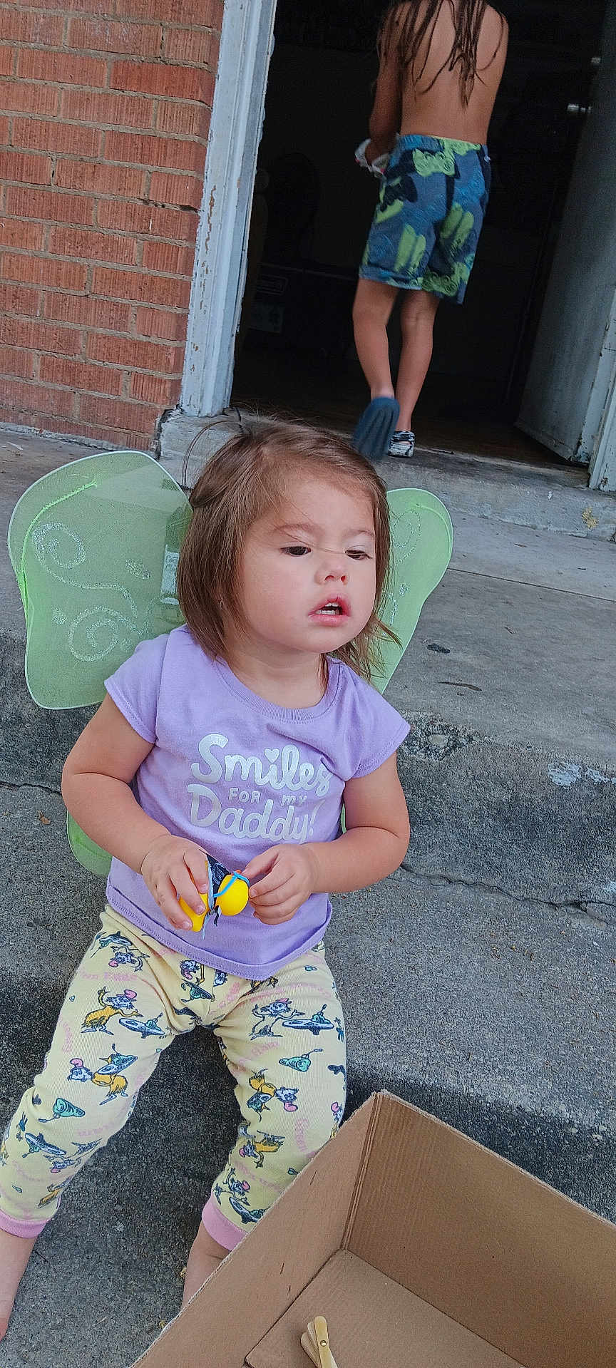 Aliveah is registered to the contest to win money with this photo: child, toddler, fairy_wings, toy, purple_shirt, pants, barefoot, concrete, doorway, brick_wall, cardboard_box, outdoor, person, playing, casual_clothing, summer, young_child, back_view, shorts, footwear