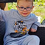 child, glasses, sweatshirt, gray_clothing, sitting, chair, outdoor, greenery, smiling, casual, footwear, playful_text, young_boy, comfort, daylight, portrait, relaxed, woven_chair, fashion, happy