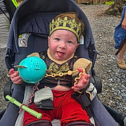 Jameson joined the competition — help win amazing prizes! baby, child, stroller, crown, toy, red_pants, outdoor, gravel, wooden_fence, tongue_out, playful, costume, seatbelt, person, cute, smiling, hand, face, headwear, casual_clothing