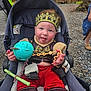 baby, child, stroller, crown, toy, red_pants, outdoor, gravel, wooden_fence, tongue_out, playful, costume, seatbelt, person, cute, smiling, hand, face, headwear, casual_clothing